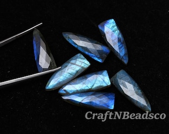 May include: A collection of faceted, elongated triangular labradorite gemstones displaying iridescent blue flashes. One stone is held by silver-toned metal tweezers. The text CraftNBeadsCo is visible in the lower right corner of the image.