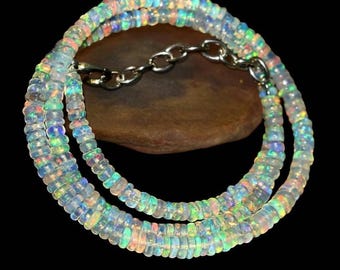 May include: A multi-strand necklace composed of small, iridescent opal beads. The beads display a range of colors, including blue, green, and orange, and are strung together with a silver chain clasp. The necklace is displayed against a dark background.