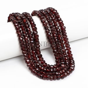May include: Strands of faceted, deep red garnet beads. The beads are strung together, creating multiple rows of the gemstone. The beads have a polished, reflective surface, and are displayed against a white background.