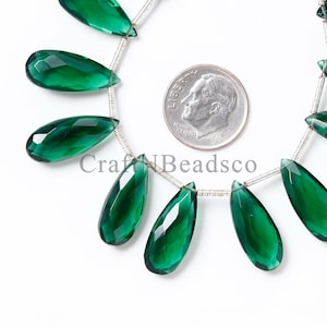 May include: Close-up of a strand of faceted, emerald green teardrop-shaped beads. A U.S. dime is placed next to the beads for size comparison. The beads are strung on a thin, silver-colored wire. The text "CraftBeadsco" is visible.