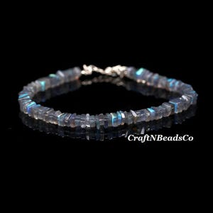 May include: A labradorite bracelet with a silver clasp. The bracelet is made of small, square-cut labradorite beads in shades of grey and blue. The beads are strung together to form a flexible band. The background is black.
