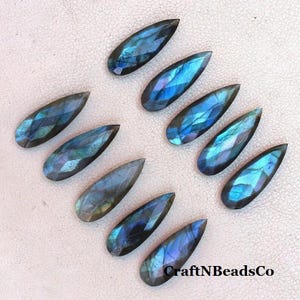 May include: A collection of ten teardrop-shaped labradorite gemstones. The stones display a range of blues, greens, and purples with a faceted cut. The gemstones are arranged on a light-colored surface. The text "CraftNBeadsCo" is visible in the bottom right corner.