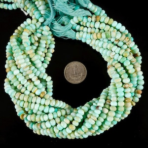 May include: Strands of light blue and tan gemstone beads. The beads are small, rounded, and strung together. A quarter is included for size reference. The beads are arranged in a circular pattern against a black background.