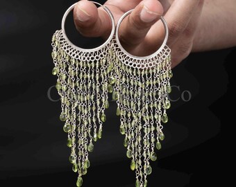 May include: A pair of silver hoop earrings featuring multiple dangling chains adorned with small, faceted light green peridot beads. The chains are attached to the bottom half of each circular hoop, creating a cascading fringe effect with teardrop-shaped stones.