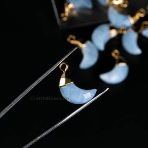 May include: A close-up of a light blue crescent moon charm with a gold-toned bail. The charm is held by a pair of tweezers against a black background.