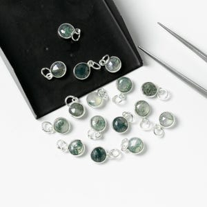 Puede incluir: Una colección de 15 dijes de plata con piedras de ágata verde musgo. Los dijes están dispuestos sobre una superficie blanca y una bandeja negra.