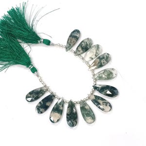 Puede incluir: Un collar de cuentas de ágata musgosa en forma de lágrima con un corte facetado. Las cuentas son una mezcla de verde, blanco y transparente, ensartadas con pequeñas cuentas de color plateado. Dos borlas verdes están unidas al collar.