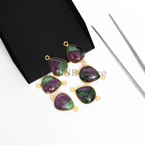 May include: Six teardrop-shaped gemstone pendants with gold-coloured frames. The gemstones feature a mix of deep purple and green hues. The pendants are arranged on a white surface with a black box in the background.