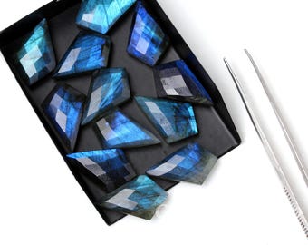 May include: A collection of eleven faceted labradorite gemstones with iridescent blue flashes arranged in a black rectangular sorting tray, accompanied by a pair of silver metal tweezers positioned to the right of the tray.