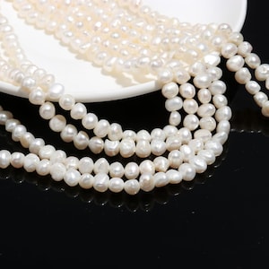 Könnte beinhalten: Eine Strähne weißer Süßwasserperlen mit leicht unregelmäßiger Form, die lose auf einer weißen Oberfläche angeordnet sind.