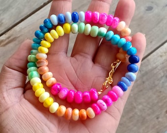 May include: A colorful beaded necklace with a gold-colored clasp. The necklace features a rainbow of colors, including pink, orange, yellow, green, blue, and white beads. The beads are small and round, and the necklace appears to be handmade.