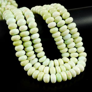 May include: A close-up of several strands of pale green gemstone beads. The beads are oval-shaped and strung together, creating a layered effect. The beads have a smooth, polished surface and a subtle, natural variation in color.