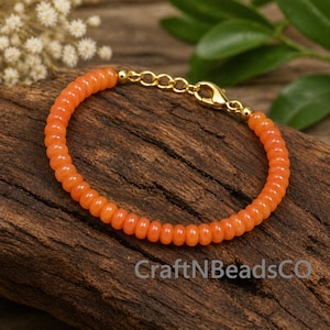Könnte beinhalten: Orangenes Perlenarmband mit goldfarbenem Verschluss und Kette. Das Armband besteht aus kleinen, runden Perlen und wird auf einer Holzoberfläche präsentiert. Der Text "CraftNBeadsCO" ist auf dem Holz zu sehen.