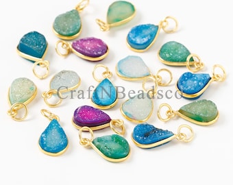 May include: A collection of teardrop-shaped druzy charms in various colors including blue, green, and purple, each encased in a gold-toned metal bezel with a small circular jump ring attached at the top for jewelry making purposes.