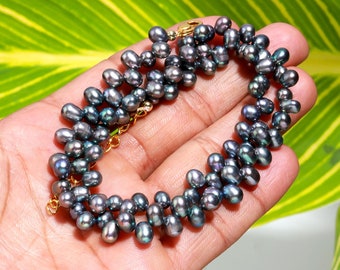 May include: A dark grey pearl bracelet with a gold clasp and chain. The pearls are clustered, creating a textured look. The bracelet is shown against a green leaf background.
