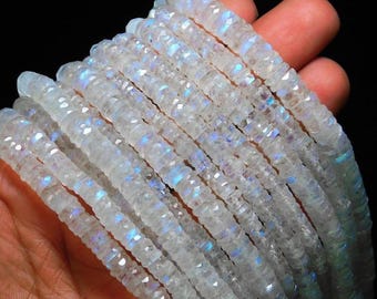 May include: Strands of iridescent white gemstone beads. The beads have a faceted, rectangular shape, and display flashes of blue and purple. The beads are held in a hand against a dark background. The text "CraftNBeadsCo" is visible at the bottom.