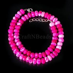 May include: A necklace featuring bright pink beads with a carved pumpkin design. The beads are strung together in a double strand. The necklace has a silver-coloured clasp and a chain extender. The text "CraftNBeads" is visible.