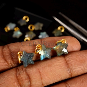 Puede incluir: Primer plano de tres colgantes de piedra preciosa de labradorita en forma de estrella con lazos dorados. Las piedras preciosas tienen una mezcla de tonos azules, verdes y grises. Hay colgantes adicionales visibles en el fondo. Los colgantes son pequeños, probablemente para hacer joyas.