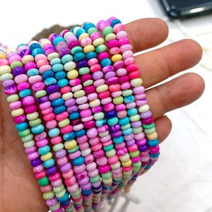 Puede incluir: Una colección de hilos de cuentas coloridas en tonos rosa, morado, azul, verde y amarillo. Las cuentas son pequeñas y redondas, y los hilos se sostienen en una mano. Las cuentas probablemente se utilizan para hacer joyas.