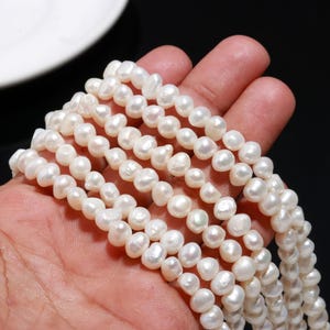 May include: Strands of white freshwater pearls. The pearls are irregular in shape and have a lustrous sheen. The pearls are strung together, creating a necklace or bracelet. The pearls are held in a hand.