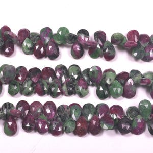 Pode incluir: Um fio de contas de pedras preciosas verdes e rosa em forma de pêra. As contas são facetadas e têm um aspecto natural e áspero.