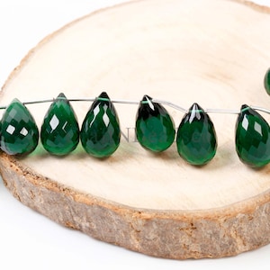 May include: A strand of faceted, teardrop-shaped green gemstones. The gems are strung on a thin, silver-colored wire and are displayed on a light brown, wooden surface. The gems have a glossy, reflective surface.