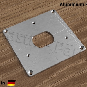 May include: A silver aluminium plate with a large circular cutout and five smaller holes. The plate has a ruler marked in centimetres along one edge. The text "Aluminium Platte" is printed in black at the top of the image. The text "made in" and a German flag are printed at the bottom of the image.