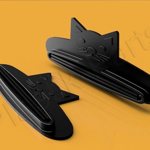 May include: Two black plastic toothbrush holders shaped like cats. The holders feature a raised design of a cat's face with whiskers and a small nose.