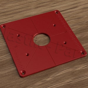 May include: A red square router plate with a large center hole and multiple smaller holes around the perimeter. The plate has a ruler on one side.