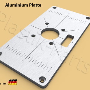 May include: A silver aluminium plate with a circular hole in the centre and a rectangular slot on the right side. The plate has a ruler on the bottom edge and a grid pattern with four holes in the corners. The text "Aluminium Platte" is printed at the top of the plate. The text "made in" and a German flag are printed at the bottom left corner of the plate.