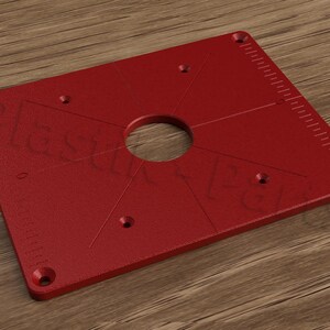 May include: Red router table insert plate with a large centre hole and five smaller holes around the perimeter. The plate has a ruler on the right side.
