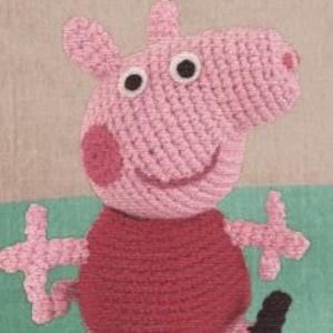 Puede incluir: Juguete de cerdo de crochet rosa con ojos negros y una camisa roja. El cerdo está de pie sobre un fondo verde y marrón.
