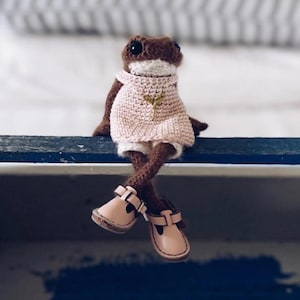Puede incluir: Una muñeca de rana de ganchillo hecha a mano con detalles marrones y blancos, que lleva un suéter rosa y zapatos marrones claros. La rana está sentada con las piernas cruzadas, mostrando los zapatos. Una pequeña hoja dorada está cosida en el suéter.