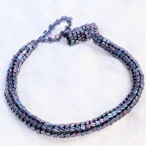 May include: A beaded necklace in shades of purple, blue, and green. The necklace is made of small, square beads woven together in a braided pattern. The clasp is a loop and bead closure. The necklace is displayed on a white, fluffy surface.