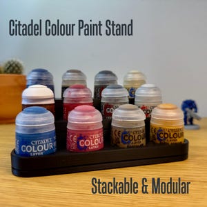 Peut inclure: Support de peinture noir à plusieurs niveaux contenant plusieurs pots de peinture Citadel Colour. Le support porte les inscriptions "Citadel Colour Paint Stand" et "Stackable & Modular". Les pots de peinture sont de différentes couleurs, dont le bleu, le rouge et le jaune.