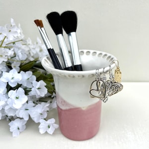 May include: A ceramic makeup brush holder with a white top and a pink base. The holder contains three makeup brushes and a small brush. Several earrings hang from the rim. White flowers are in the background.