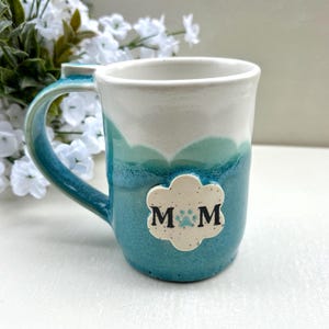 May include: A handmade ceramic mug with a blue and white gradient design. The mug features a large handle and a decorative cloud-shaped emblem with the letters "MOM" and a paw print. The mug is a unique gift for pet lovers.