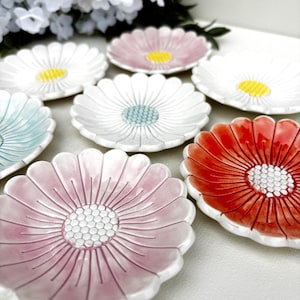 May include: A collection of ceramic plates shaped like flowers with scalloped petals. The plates feature various colors including pink, white, light blue, and orange, each with a textured, raised center resembling a flower head in contrasting colors.