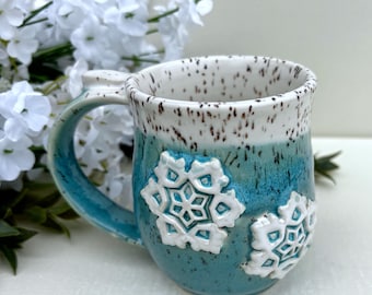 May include: A ceramic mug featuring a teal and white ombre glaze with dark brown speckles throughout. The exterior is decorated with two raised, white, embossed snowflake designs. The mug has a curved handle and a smooth, rounded rim.