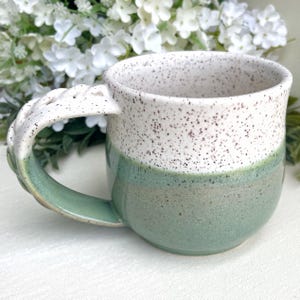 May include: A handmade ceramic mug with a unique handle design. The mug features a two-tone color scheme, with a speckled white upper half and a muted green lower half. The handle is white with a textured, openwork design.