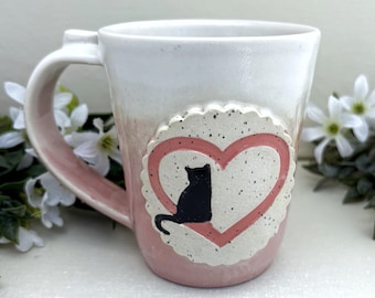 May include: A ceramic mug with a white top and pink bottom featuring a speckled, scalloped circular emblem on the front. The emblem displays a pink heart outline with a solid black cat silhouette sitting inside the heart shape on the mug surface.