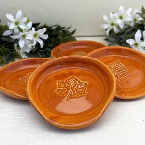 May include: Three round ceramic dishes with a glossy orange glaze. Each dish features a distinct embossed leaf pattern in the center, including a maple leaf and a fern frond. The edges are slightly raised and rounded, creating a shallow bowl shape.