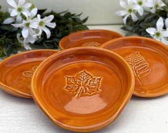 May include: Three round ceramic dishes with a glossy orange glaze. Each dish features a distinct embossed leaf pattern in the centre, including a maple leaf and a fern frond. The edges are slightly raised and rounded, creating a shallow bowl shape.
