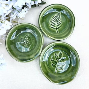 May include: Three green ceramic spoon rests with leaf designs. Each round rest features a different embossed leaf pattern: a fern, a maple leaf, and a sprig of leaves. The spoon rests are glossy and have a small lip for holding utensils.