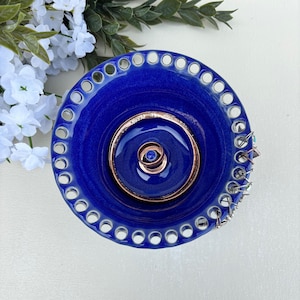 May include: A cobalt blue ceramic dish with a decorative pierced rim. The center features concentric copper-colored rings and a small blue gem. A few silver-toned jewelry pieces are placed on the side.
