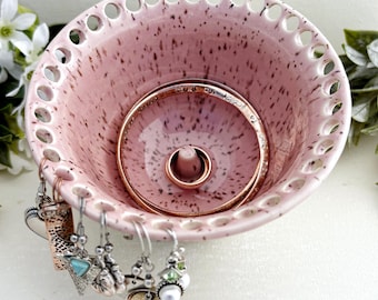 May include: A light pink ceramic jewellery bowl with dark brown speckles, featuring a perforated rim for hanging earrings and a central cone for holding rings. A metallic ring rests on the cone, and several dangling earrings hang from the rim.