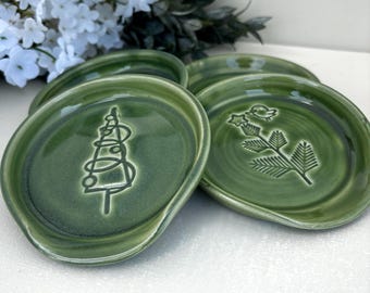 May include: A set of round, sage green ceramic spoon rests with a glossy finish. One spoon rest features an embossed line drawing of a stylised Christmas tree, while the other displays an embossed branch with a small bird and star.