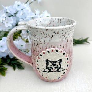 May include: A ceramic mug with a speckled pink and white glaze featuring a circular cream-colored medallion on the front. The medallion displays a black and white illustration of a cat peeking over a ledge, surrounded by small pink and black dots.