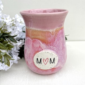 May include: A ceramic tumbler with a pink and white glazed finish featuring a central oval plaque with speckles. The plaque displays the text M, a pink heart, and M in black lettering. The vessel has a flared rim and a rounded base.