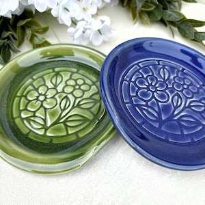 Può includere: Due poggia cucchiaio rotondi in ceramica, uno in verde oliva lucido e l'altro in blu reale profondo. Ogni pezzo presenta un design centrale in rilievo di un fiore con foglie, racchiuso in un bordo circolare, con una finitura smaltata liscia.